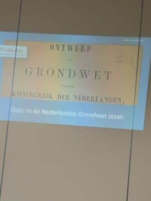 Een geprojecteerde dia met het Ontwerp van Grondwet van het Koninkrijk der Nederlanden en de vraag: in de Nederlandse Grondwet staat.