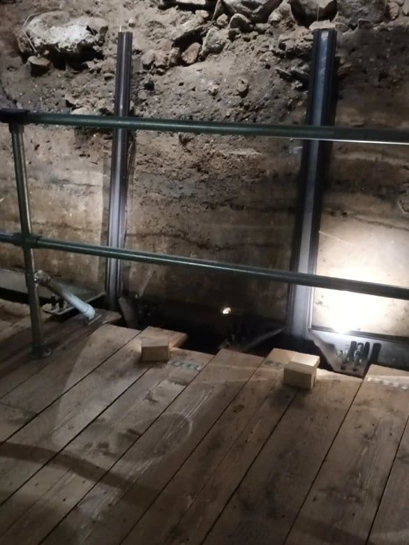 Een opgraving achter glas: muurresten en aardlagen onder de grond, met een houten vloer ervoor.