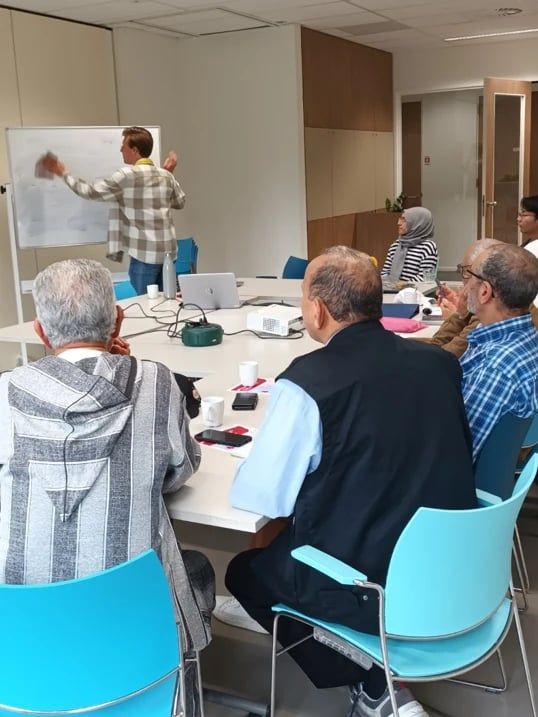 Deelnemers aan tafel luisteren naar een docent die iets op een whiteboard schrijft.